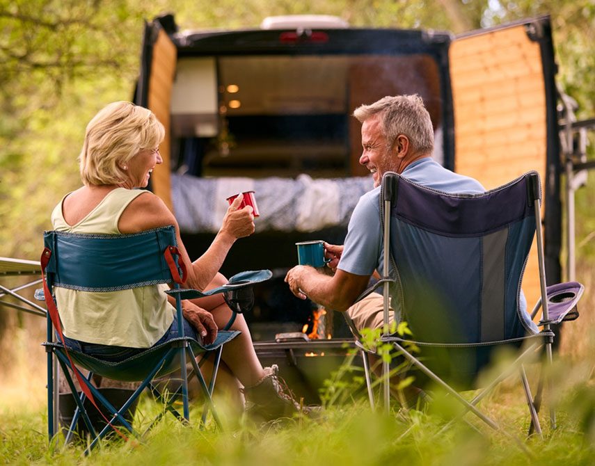Rear View Of Senior Couple Camping In Countryside With RV Drinking Coffee By Outdoor Fire